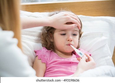 Mother Measuring Temperature Of Her Ill Kid. Sick Child With High Fever Laying In Bed And Mother Holding Thermometer. Hand On Forehead. 