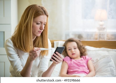 Mother Measuring Temperature Of Her Ill Kid. Sick Child With High Fever Laying In Bed And Mother Holding Thermometer. Mother With Cell Phone Calling To Doctor