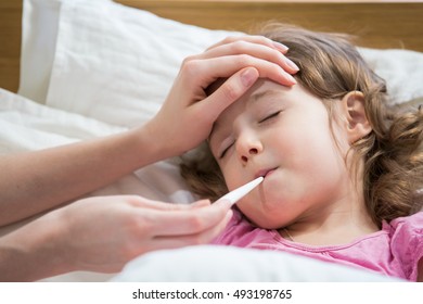 Mother Measuring Temperature Of Her Ill Kid. Sick Child With High Fever Laying In Bed And Mother Holding Thermometer. Hand On Forehead. 