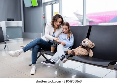 Mother With Paper Cup And Daughter Looking At Smartphone In Lounge Hall Of Airport