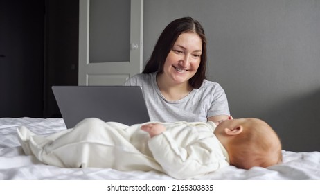 Mother Pays Attention To Little Baby Girl Sitting Near Bed And Working On Laptop. Newborn Child Tries To Draw Attention Of Mom Lying On Parent Bed