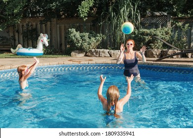 Mother Playing Ball With Daughters Children In Swimming Pool On Home Backyard. Mom And Sisters Siblings Having Fun In Swimming Pool Together. Summer Outdoor Water Activity For Family And Kids. 