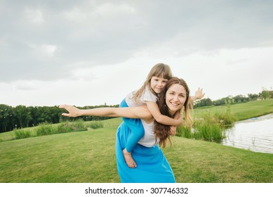 Mother Playing With Daughter Outdoor. Girl On A Piggyback, Mom Doing Airplane With Joy 