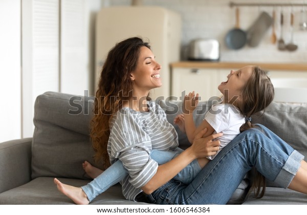 Mother playing with little daughter, mom enjoy\
time on weekend with pre-school cheery kid girl tickles her\
laughing together seated on couch at home, having fun, activities\
with child at home\
concept