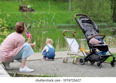 Mother playing with little daughter in a park near the pond. Woman blows soap bubbles, family leisure