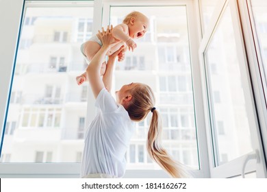 Mother Playing With Newborn Baby Son At Home. Woman Tossing Kid Up On Balcony. Happy Infant Wearing Diaper