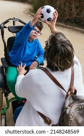 A Mother Playing With A Soccer Ball With Her Son With A Disability In A Wheelchair In The Park.