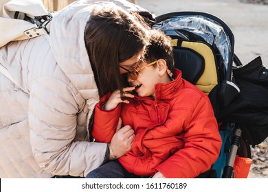 A Mother Plays With Her Son With Multiple Disabilities In A Park, The Child Goes With A Wheelchair, It Is Winter And They Go With Warm Clothes. The Woman Sticks Out Her Tongue, Tickles And Laughs Play