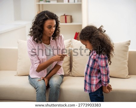 Mother pointing finger while talking to child standing with downcast posture in living room.