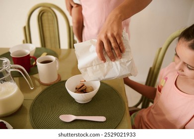 A mother pours cereal into a bowl for her child at the breakfast table, creating a warm family morning routine with milk and coffee. - Powered by Shutterstock