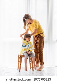 Mother Prepares To Cut Her Son's Hair By Herself. Little Boy Sits, Covered With Cloth, And Holds Pair Of Scissors. New Normal In Quarantine Of Coronavirus COVID-19. Self Care At Home Lifestyle.