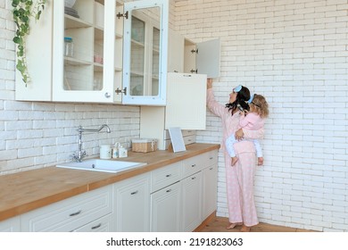 Mother Prepearing Breakfast Together With Dauther At Home In Kitchen. Toddler Watching Mobile Phone. Smiling Young Caucasian Woman In Pajama Sleep Mask Cooking Food Indoors With Girl Child On Her Arms