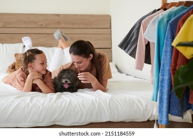 Mother And Preteen Daughter Chatting When Resting On Bed With Small Dog, Considered Family Member