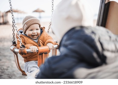 Mother pushing her cheerful infant baby boy child on a swing on sandy beach playground outdoors on nice sunny cold winter day in Malaga, Spain - Powered by Shutterstock