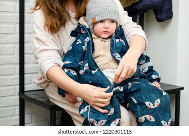 Mother Puts A Blue Overalls On The Toddler Baby Sitting In The Home Hallway. Woman Mom Dressing Warm Clothes Coverall On Child For Winter Walk In Cold Weather. Kid Aged One Year And Three Months