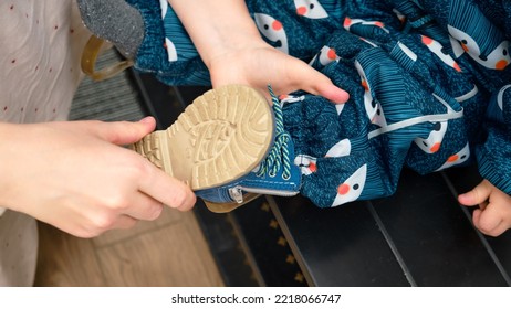 The Mother Puts A Blue Shoes On The Foot Toddler Baby Sitting In The Home Hallway. Woman Mom Dressing Warm Boots Clothes On Child For Winter Walk In Cold Weather. Kid Aged One Year And Three Months