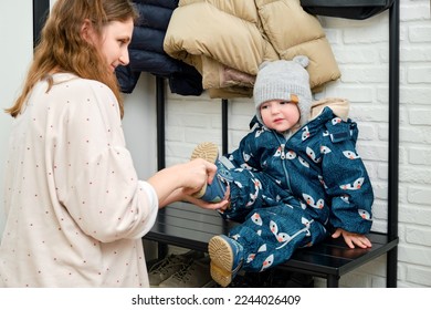 The mother puts a blue shoes on the foot toddler baby sitting in the home hallway. Woman mom dressing warm boots clothes on child for winter walk in cold weather. Kid aged one year and three months