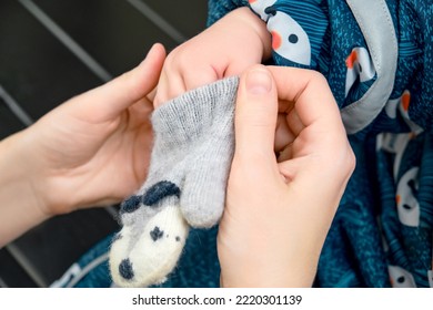 Mother Puts A Grey Mitten On The Hand Toddler Baby Sitting In The Home Hallway. Woman Mom Dressing Warm Clothes Glove On Child Hand For Winter Walk In Cold Weather. Kid Aged One Year And Three Months