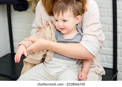 A Mother Puts A Hoodie Jacket On A Toddler Baby Sitting In The Home Hallway. Woman Mom Dressing Warm Clothes On Child For Winter Walk In Cold Weather. Kid Aged One Year And Three Months