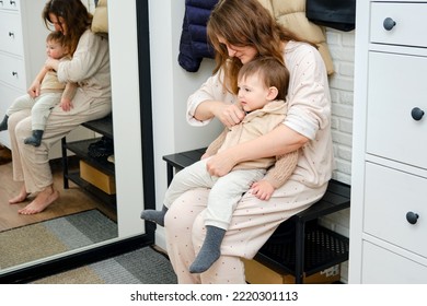 A Mother Puts A Hoodie Jacket On A Toddler Baby Sitting In The Home Hallway. Woman Mom Dressing Warm Clothes On Child For Winter Walk In Cold Weather. Kid Aged One Year And Three Months