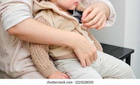 A Mother Puts A Hoodie Jacket On A Toddler Baby Sitting In The Home Hallway. Woman Mom Dressing Warm Clothes On Child For Winter Walk In Cold Weather. Kid Aged One Year And Three Months