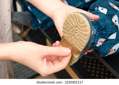 The Mother Puts A Strip For Shoes On The Foot Toddler Baby Sitting In The Home Hallway. Woman Mom Dressing Warm Boots On Child For Winter Walk In Cold Weather. Kid Aged One Year And Three Months