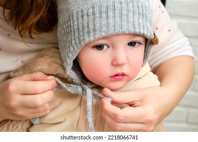 The Mother Puts A Warm Hat On The Head Toddler Baby Sitting In The Home Hallway. Woman Mom Dressing Warm Clothes On Child For Winter Walk In Cold Weather. Kid Aged One Year And Three Months