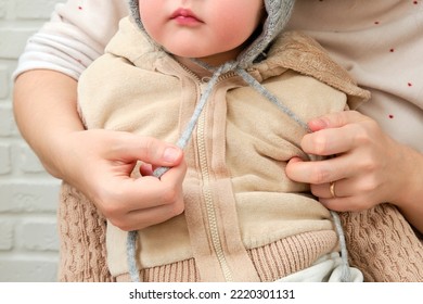 The Mother Puts A Warm Hat On The Head Toddler Baby Sitting In The Home Hallway. Woman Mom Dressing Warm Clothes On Child For Winter Walk In Cold Weather. Kid Aged One Year And Three Months