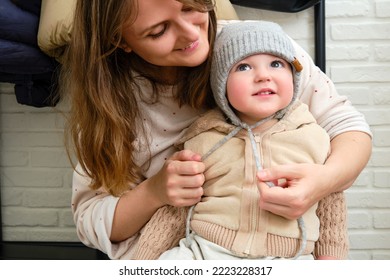 The Mother Puts A Warm Hat On The Head Toddler Baby Sitting In The Home Hallway. Woman Mom Dressing Warm Clothes On Child For Winter Walk In Cold Weather. Kid Aged One Year And Three Months