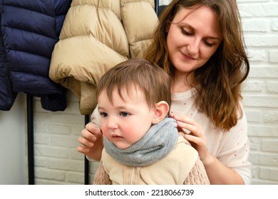 Mother Puts A Warm Scarf On The Toddler Baby Sitting In The Home Hallway. Woman Mom Dressing Warm Clothes On Child For Winter Walk In Cold Weather. Kid Aged One Year And Three Months