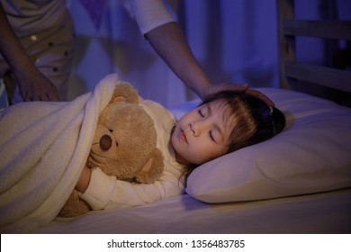 Mother putting blanket on and stroking head for her little girl daughter on bed in a dark bedroom at night, Child asian girl hug teddy bear, Comfortable children at home concept