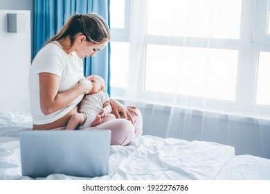 Mother In Pyjama Breastfeeding Baby Girl While Sitting On Large Bed With Grey Laptop On Foreground Under Bright Light At Home.