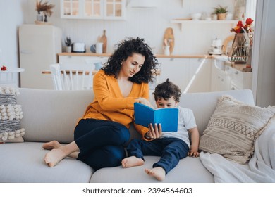 Mother reading book with son sitting on couch at home. Domestic education, childhood. Young mother entertains little boy on weekend. Self isolation, pandemic, stay home. - Powered by Shutterstock