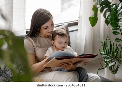 Mother reads a book to a cute little girl, sitting cozy near big window at home