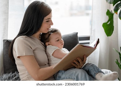 Mother reads a book to a cute little girl, sitting cozy near big window at home