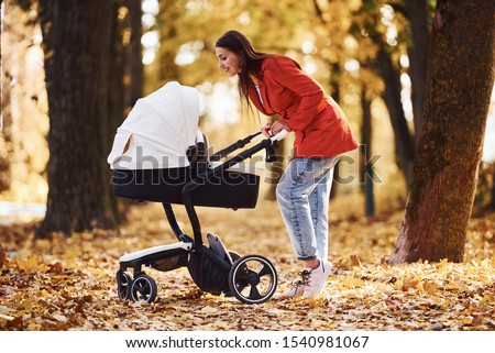 Mother in red coat have a walk with her kid in the pram in the park at autumn time.