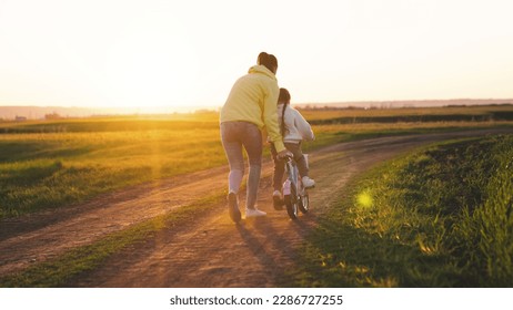 mother rides child bicycle sunset. happy family park. Mom teaches her daughter ride bike. concept happy family summer park. fun vacation. teamwork. mother runs green grass rolling child bicycle. - Powered by Shutterstock