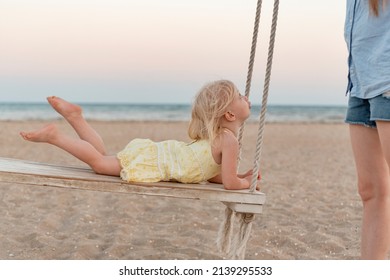 Mother rides her little daughter on swing on the seashore. Cute towheaded girl on rope swing on the beach on sea background