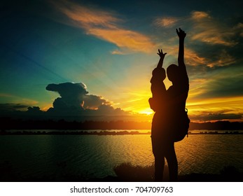 Mother Shoulder Bag Carrying Baby Standing By The Pond. They Raise Their Hands. While The Sun Is Setting.silhouette