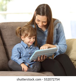Mother showing media content on line to her two years daughter in a tablet sitting on a couch in the living room in a house interior