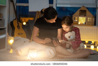 Mother Showing Phone To Little Daughter Sitting On Floor In Playroom At Night