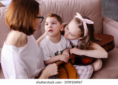 Mother And Sibling Children Play Together. A Child With Cerebral Palsy With Family In Rehabilitation. Inclusion Friendship With  Disabled Person. Music Therapy Playing The Guitar. Happy Kids Laugh Fun