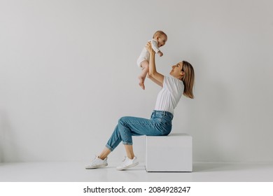 Mother Sitting And Holding Baby Up In The Air, Smiling, On White Wall Background.