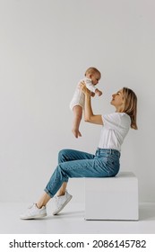 Mother Sitting And Holding Baby Up In The Air, Smiling And Playing, On White Wall Background.