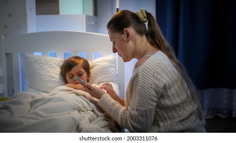 Mother Sitting Next To Sick Son In Bead And Typing Doctor Phone Number On Smartphone. Concept Of Children Illness, Disease And Parent Care.