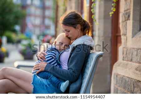 Mother, sitting on a bench in the center of beautifull old village and holding her baby boy, smiling and hugging