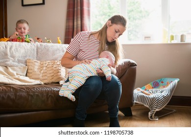 Mother Sitting On The Edge Of The Sofa In The Living Room And Burping Her Newborn Child After Feeding Him. Her Other Son Is Stadning Behind The Sofa Playing While His Toys.