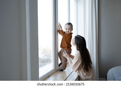 A Mother And A Small Child Stand At A Large White Window And Look Out Into The Street. Safety For Small Children In The House.