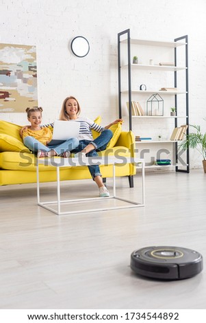 Mother smiling, hugging daughter with laptop on sofa near coffee table and looking at robotic vacuum cleaner on floor in living room Stock photo © 