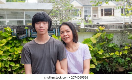 Mother And Son, Asian Teen, Black Hair. Mother And Child Concept, Hugging Each Other, Smiling Happily Looking At The Camera. At The Front Garden